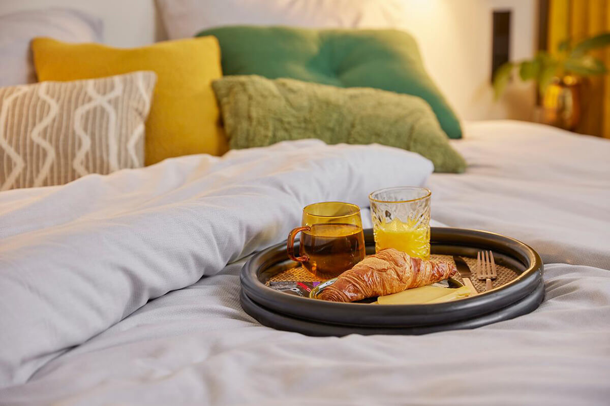 Ontbijtblad met croissants op een bed met grijs bedlinnen en gekleurde kussens